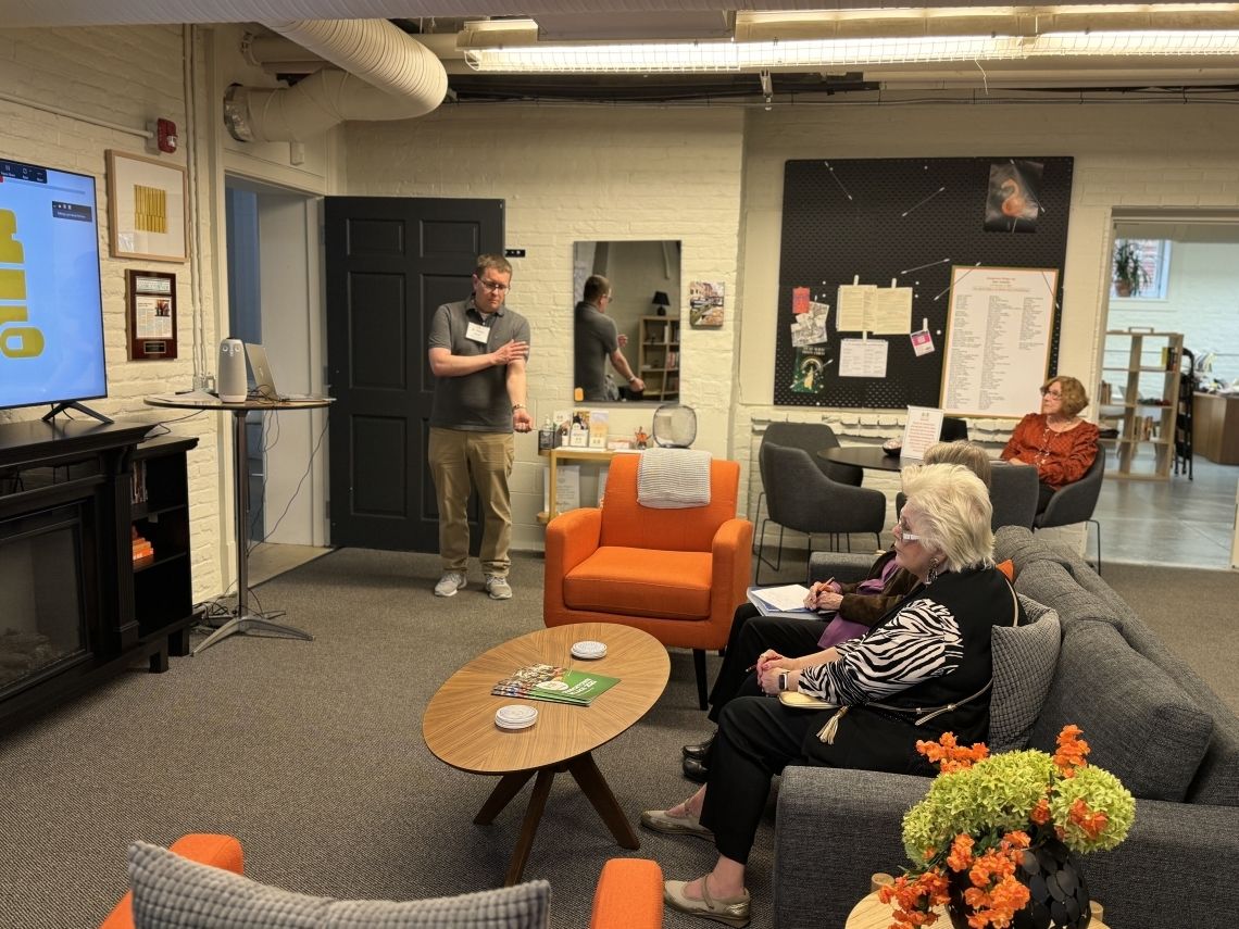 Dr. Hood stands in front of a room explaining Virtual Medical Devices to two participants at Georgetown Village seated on grey couch.