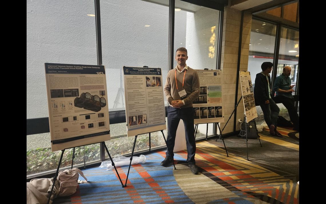 The poster presentation room at the ISMAR conference. HealthDesk representative pictured smiling with the HealthDesk poster, situated in the middle of two other poster presentations. 