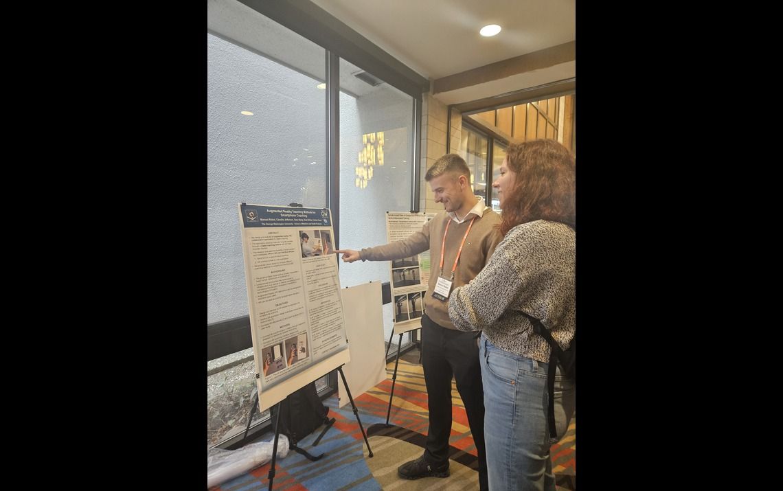 HealthDesk representative pointing to the poster, explaining the presentation to an onlooking attendee at ISMAR conference 2024.