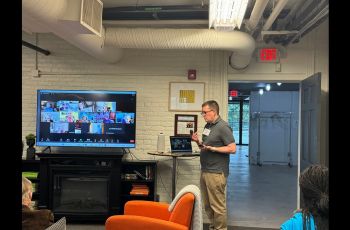 Dr. Hood giving a talk at Georgetown Village, open room with a television screen at the front displaying a Zoom screen with the virtual participants.
